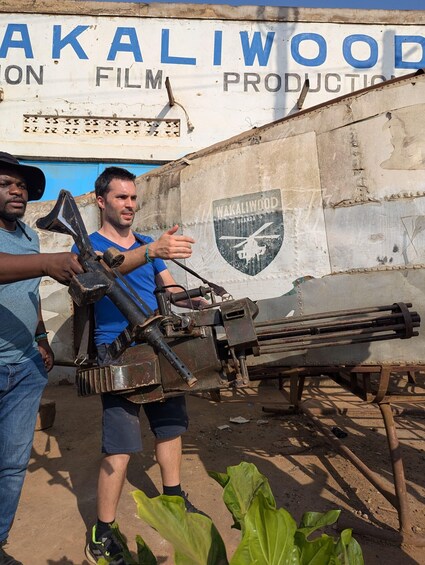 Two people holding a large, old-fashioned camera-like device in front of a building with a sign that reads 'WAKALIWOOD FILM PRODUCTION.' 5