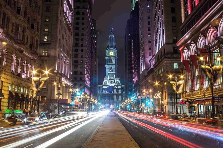 A nighttime city street with tall buildings, a prominent clock tower, and light trails from moving vehicles. 1