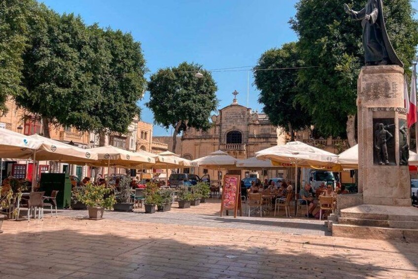 An outdoor café area with tables and umbrellas, a statue, and a building with a cross on top in the background. 4