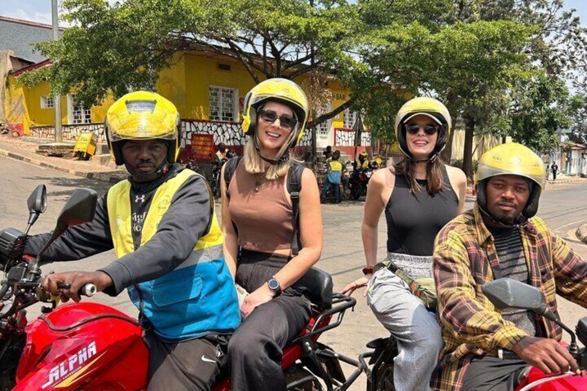 Four people on motorcycles, wearing helmets and reflective vests, on a street with buildings and trees in the background. 1