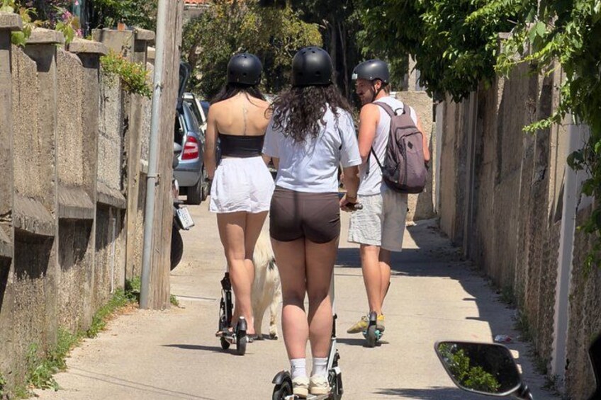 A narrow paved path with people riding scooters, a parked car, and a wall on one side. 5