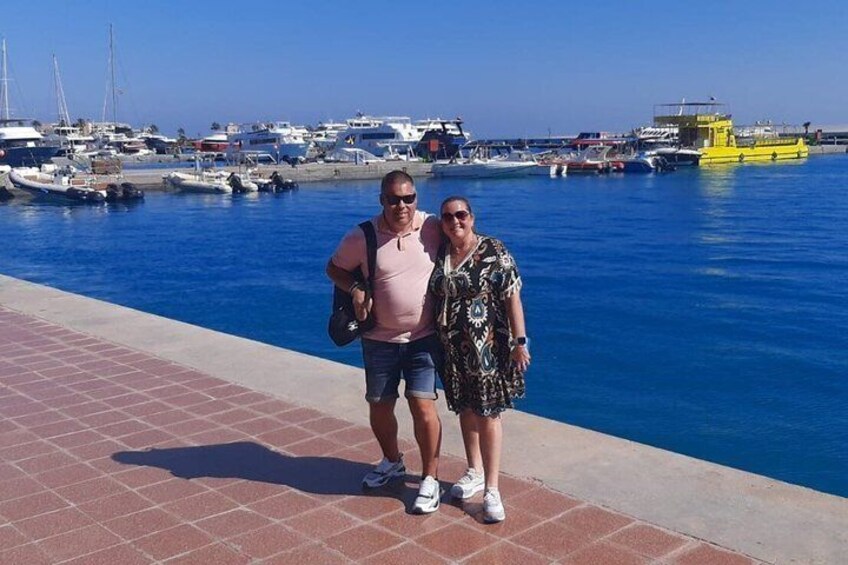 Two people standing on a tiled walkway by a marina with boats in the background. 1