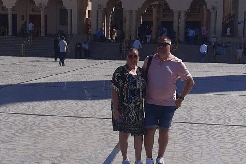 A man and a woman standing in a public square with patterned pavement and classical architecture in the background. 2