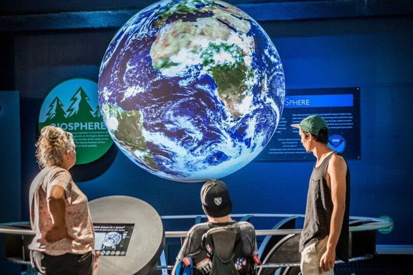 Trois personnes observent un grand globe terrestre, avec une pancarte indiquant « Biosphère » et, au fond, une autre pancarte indiquant « Sphère ». 1