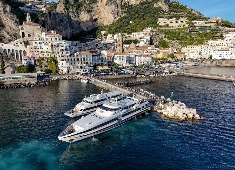 A large white yacht docked at a pier with a rocky breakwater. In the background, there are colorful buildings along the coastline and a hill with more buildings. 1