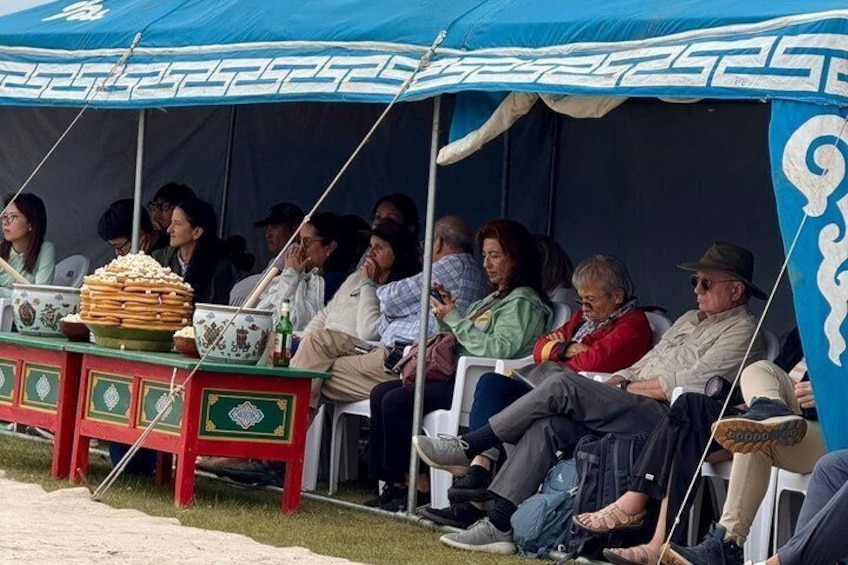 People seated under a tent with a table of food items. 2