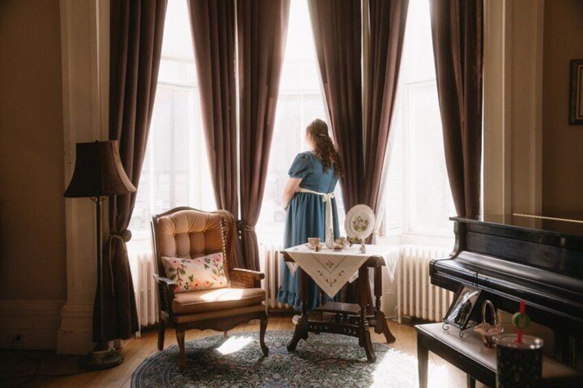 A woman in a blue dress standing by a window with curtains, a piano, and a table with a teapot and cups. 5