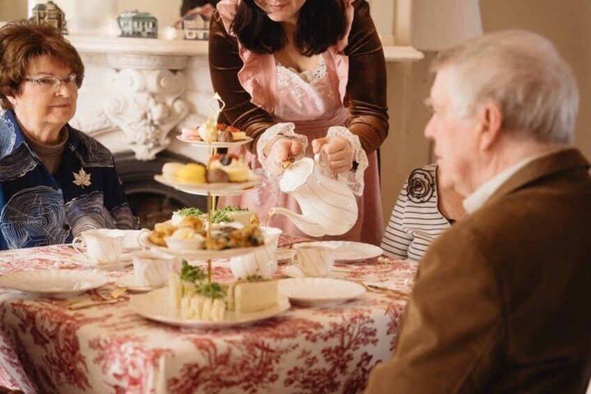 A group of people at a tea service with a tiered cake and teapots on a table. 2