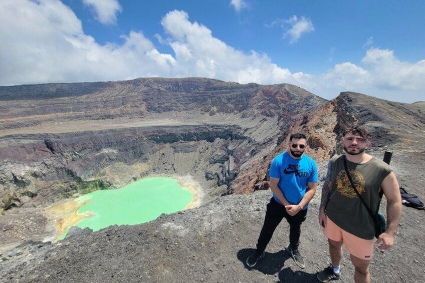 Two people standing on a rocky outcrop overlooking a green crater lake with a barren landscape in the background. 4
