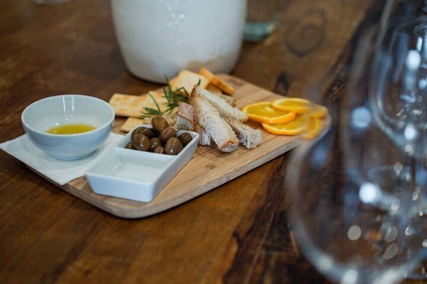 A wooden board with bread, olives, and orange slices, a bowl of olive oil, and a glass pitcher on a wooden table. 3