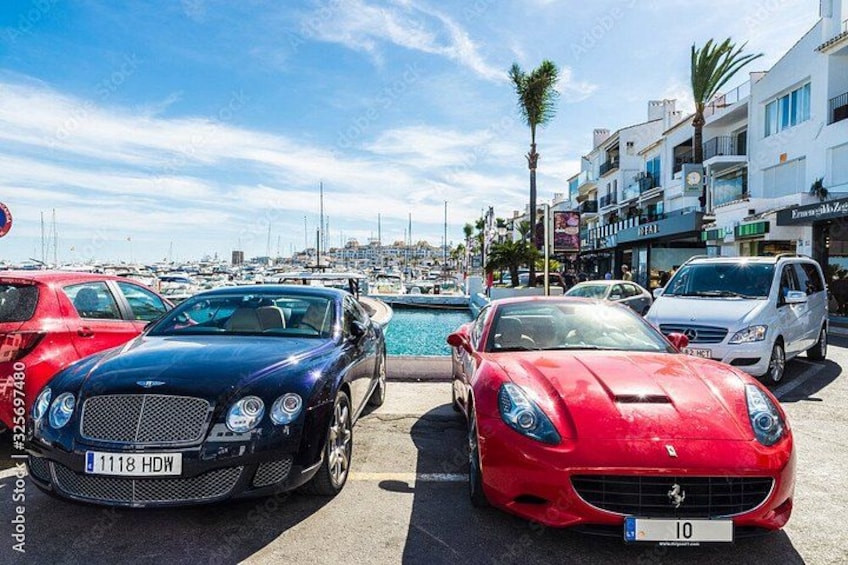 Luxury cars parked by a waterfront with a marina and buildings in the background. 5