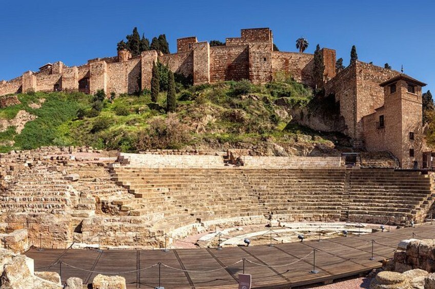 An ancient amphitheater with stone steps and a large open area, surrounded by ruins and a hilltop structure. 4