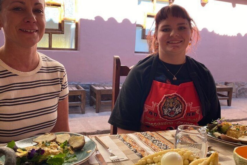 Two people seated at an outdoor table with a meal, a window, and a wall in the background. 4