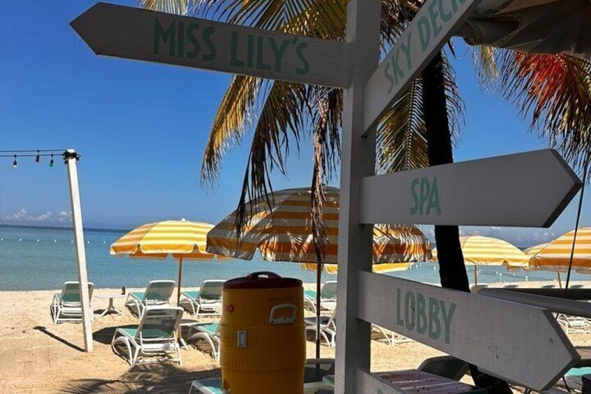 A beach with lounge chairs, umbrellas, and directional signs pointing to Miss Lily's, Sky Deck, Spa, and Lobby. 5