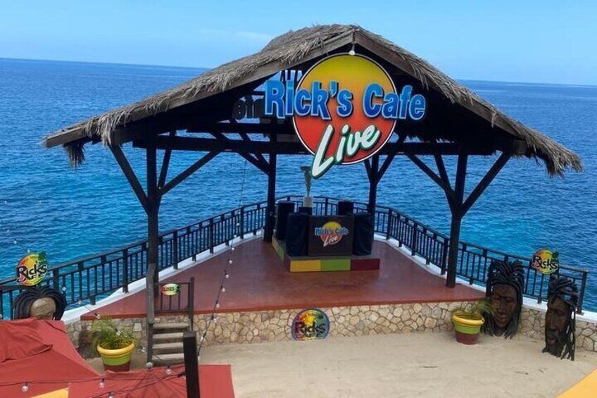 A beachside stage with a sign reading 'Rick's Cafe Live' and a thatched roof structure. 1