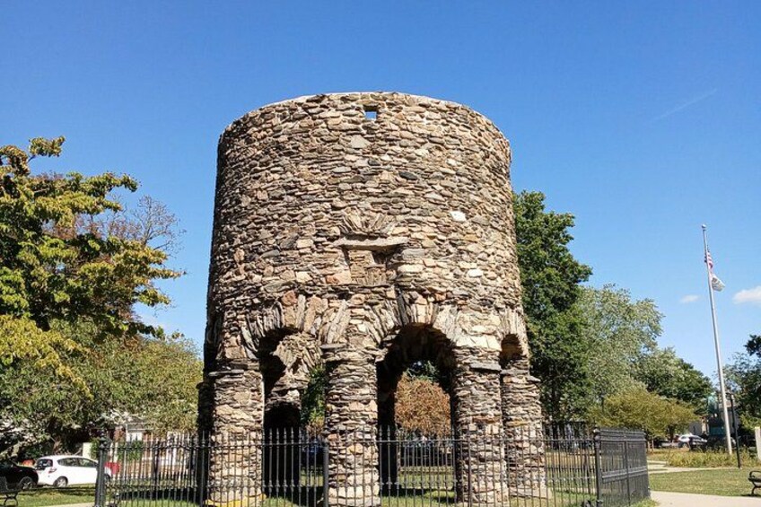 A stone tower with arches, surrounded by a fence, trees, and a clear sky. 4