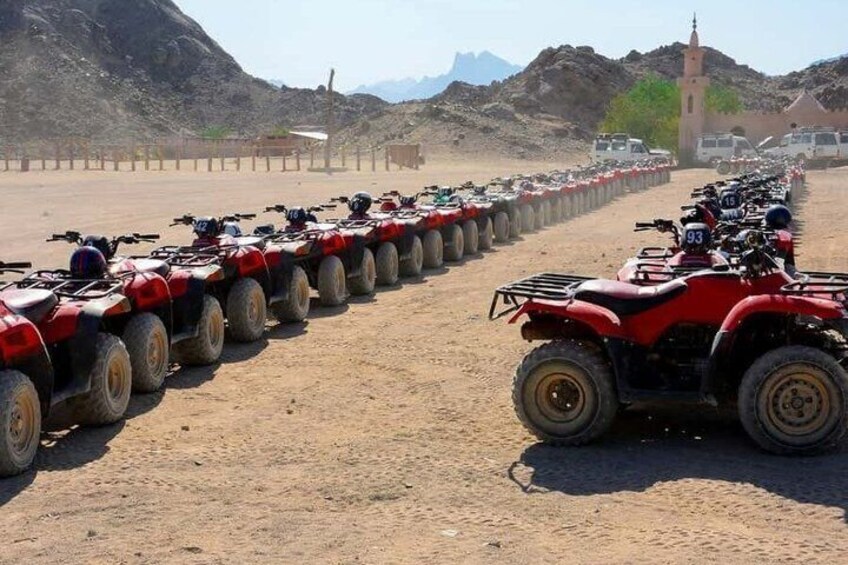 A row of red ATVs parked on a dirt road with a mountainous backdrop and a church tower. 4