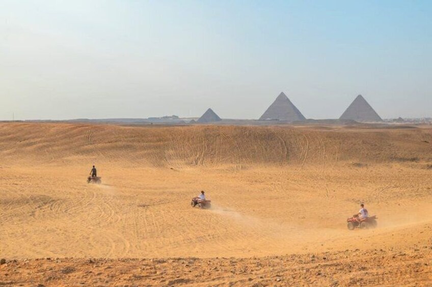 A desert landscape with three pyramids in the background and people riding off-road vehicles. 4