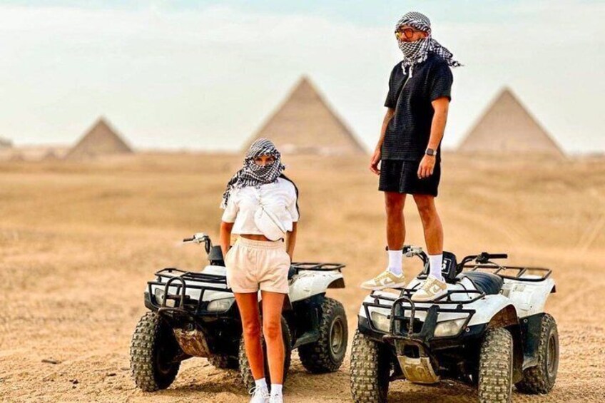 Two people on ATVs in a desert with pyramids in the background. 1