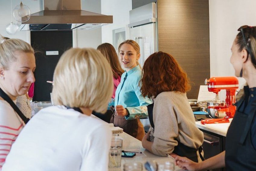 A group of people in a kitchen, some standing and some sitting, with a red mixer on the counter. 5