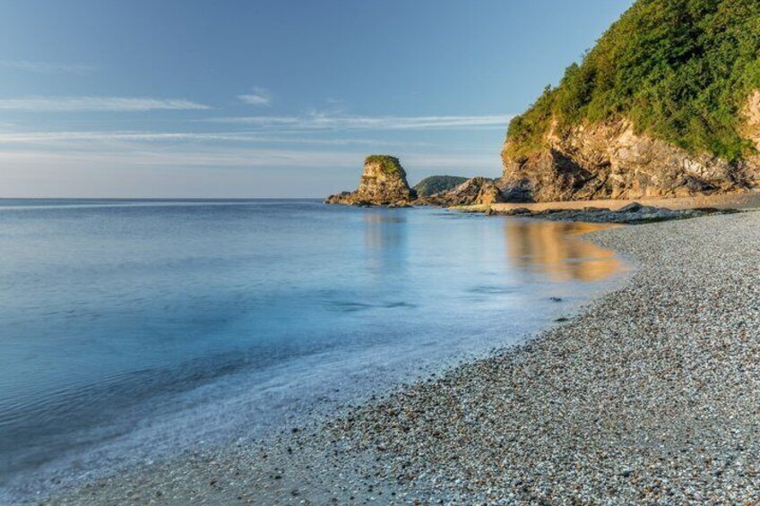 A pebbly beach with calm water, a rocky outcrop, and a green cliff. 4