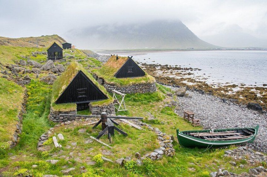 Traditional grass-roofed houses, a wooden boat, and a rocky shoreline with a mountain in the background. 3