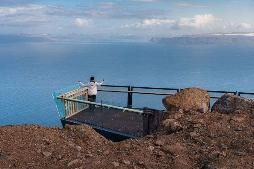 A person standing on a glass platform overlooking a body of water with a rocky landscape in the foreground. 1