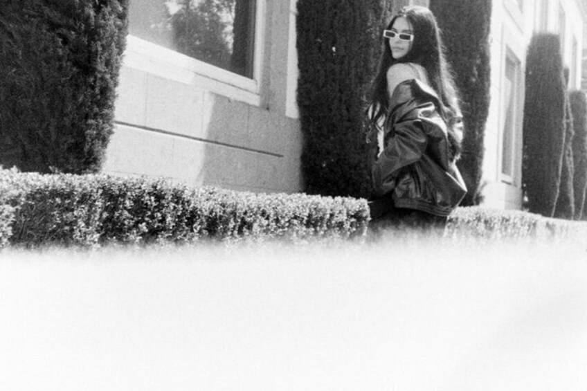 A person standing in a garden with trimmed hedges and a building in the background. 1