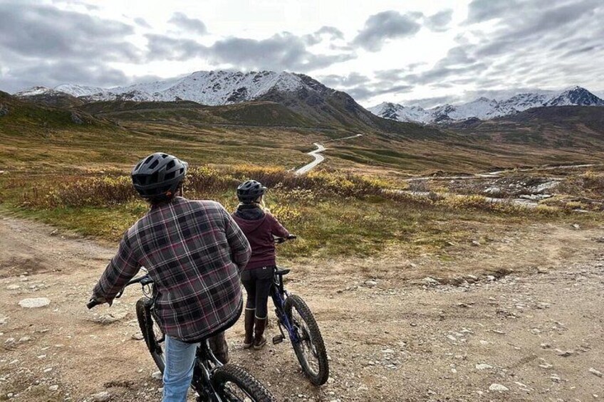 Two people on bicycles on a dirt path with mountains in the background. 2