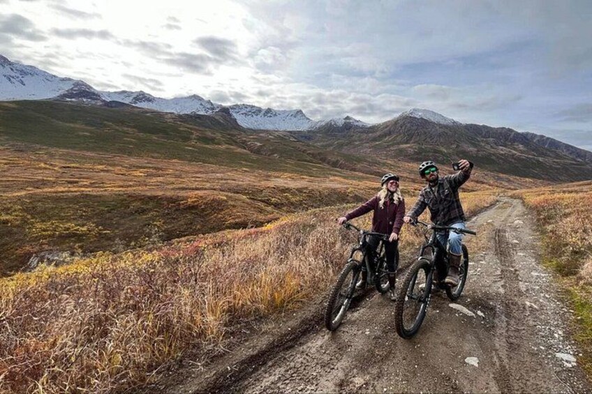 Two people on mountain bikes on a dirt path with a mountainous landscape in the background. 3