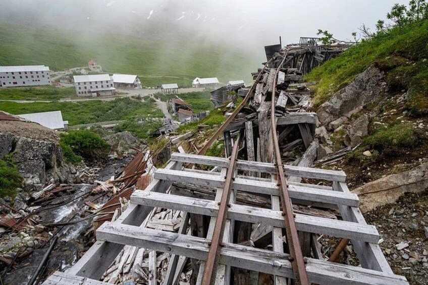 A dilapidated railway track with wooden sleepers and metal rails, surrounded by rocky terrain and buildings in the background. 5