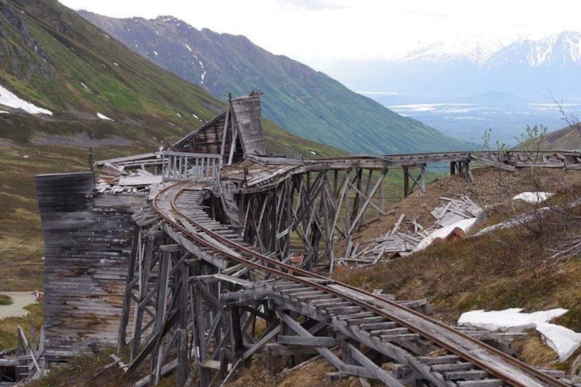 A dilapidated wooden railway bridge in a mountainous area with snow patches and a distant view of mountains. 1