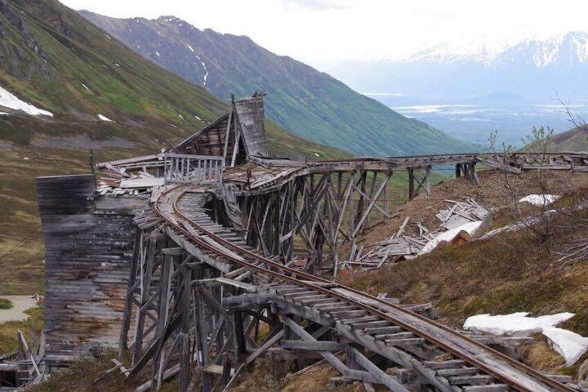 A dilapidated wooden railway bridge in a mountainous area with snow patches and a distant valley. 1