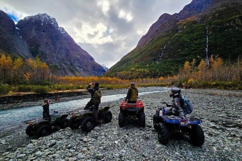 Three off-road vehicles parked on a rocky riverbank with mountains in the background. 3