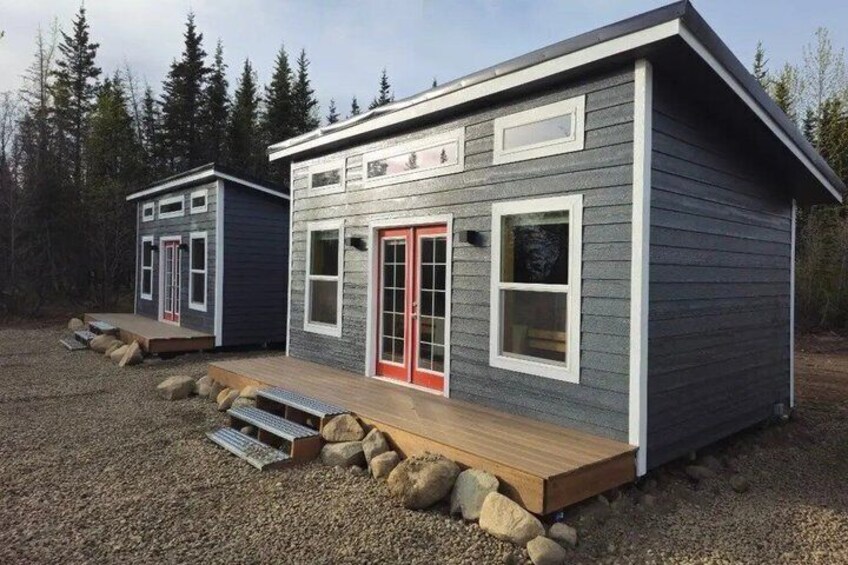 Two modern cabins with grey siding and red accents, set in a forested area with gravel ground. 5