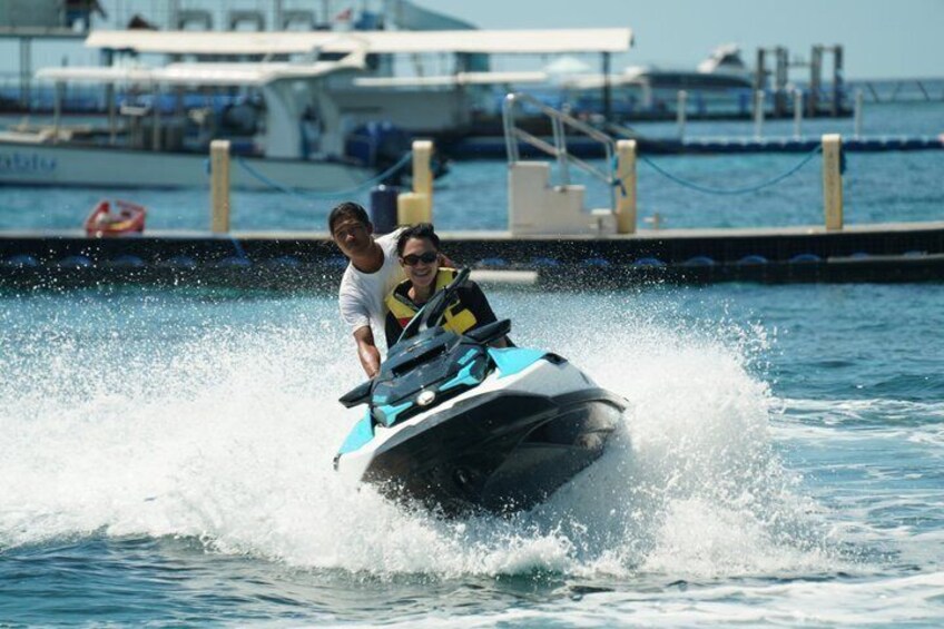A jet ski with two people, water splashing around, a dock, and boats in the background. 1
