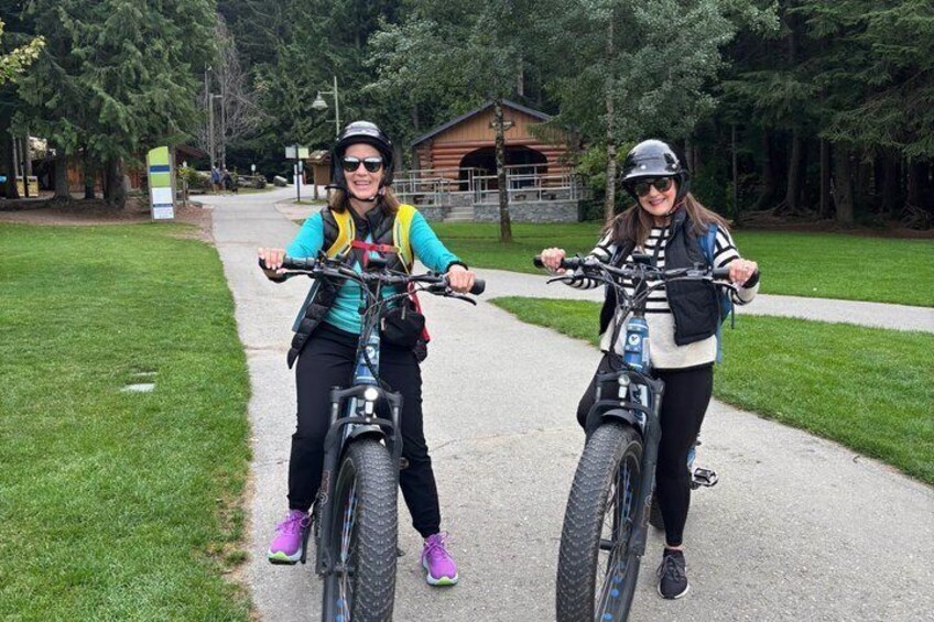 Two people on bicycles on a paved path with grass on either side, a wooden building in the background, and trees surrounding the area. 3