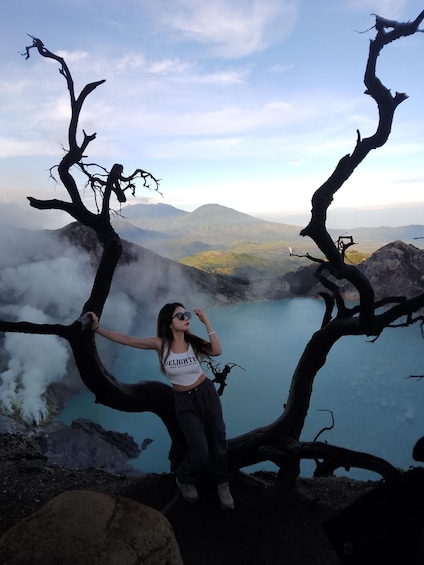 A person standing between two dead tree branches with a mountainous landscape and a lake in the background. 5