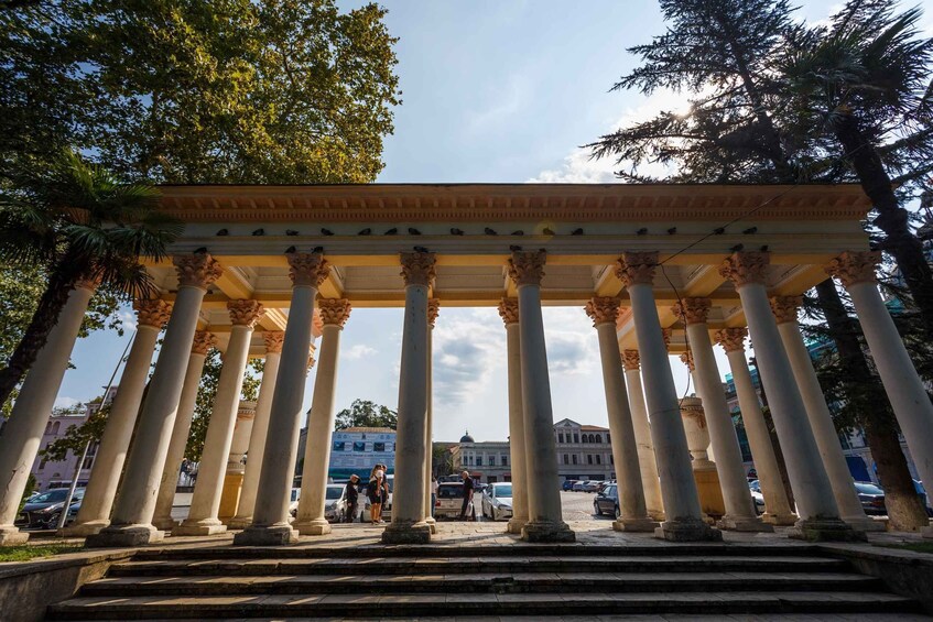 A classical structure with columns and a pediment, surrounded by trees and urban buildings. 2