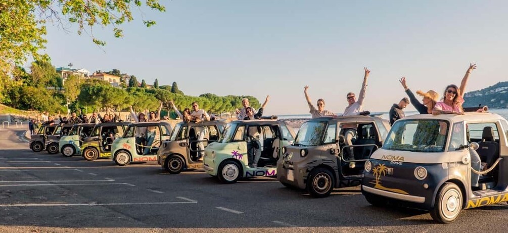 A convoy of NOMA cars with passengers raising their hands, parked on a road with a hilly landscape in the background. 1