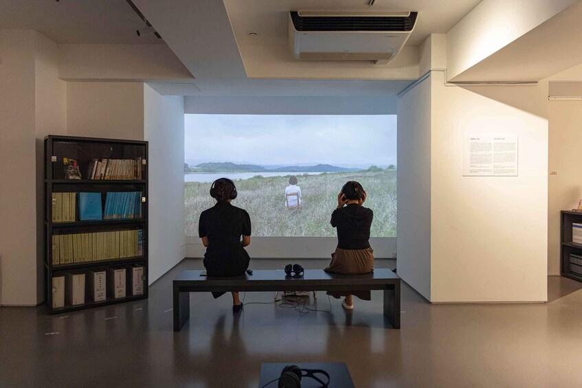 Two people sitting on a bench facing a projection screen, with a bookshelf to the left. 5