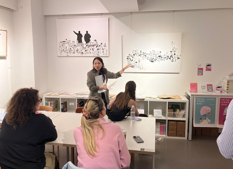A person presenting in a gallery with artwork on the walls and attendees seated at tables. 1