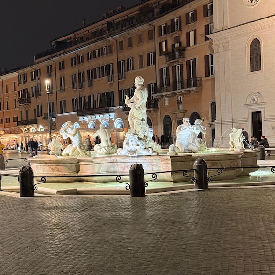 A nighttime view of a historic fountain with classical statues, surrounded by cobblestone pavement and traditional buildings. 1