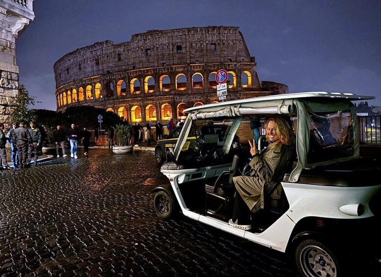 A white car parked on cobblestone pavement with the Colosseum illuminated in the background. 1
