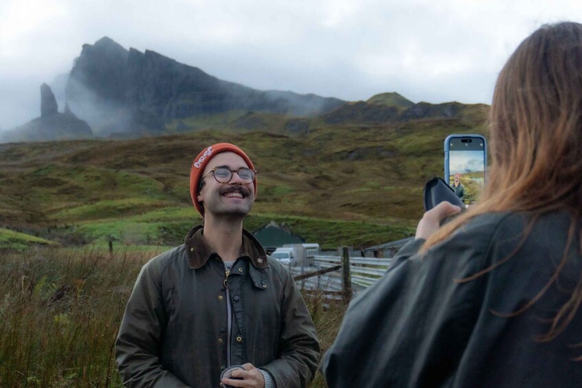 A person in a green jacket and red beanie standing in a grassy field with a mountainous landscape in the background. 4