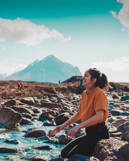 A person sitting on rocks by a river with a mountain in the background. 1