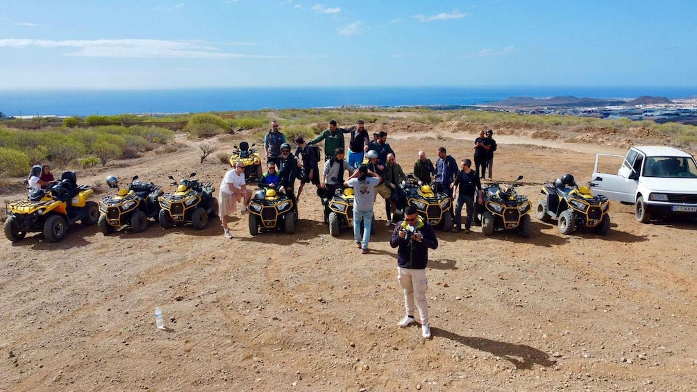 A group of people with ATVs on a dirt road with a view of the ocean in the background. 1