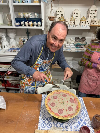A person cutting a pizza on a table in a kitchen with ceramic decorations on shelves. 3