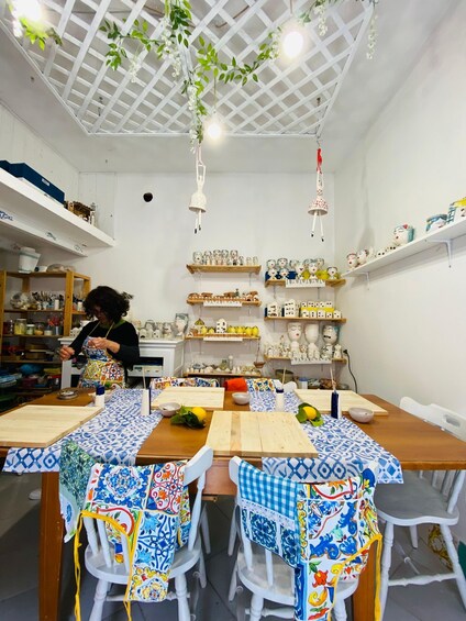 A craft workshop with tables covered in patterned cloths, shelves with ceramic figurines, and hanging plants. 1