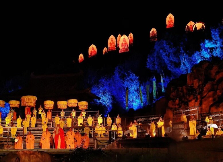 A nighttime outdoor scene with illuminated statues and figures in traditional attire, set against a backdrop of rocky terrain and illuminated trees. 5